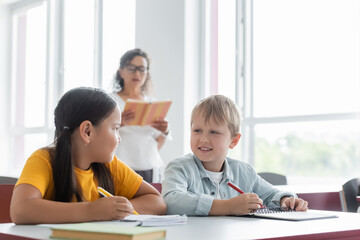 interracial pupils looking at each other while writing dictation near blurred african american...