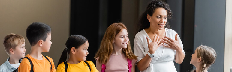 african american teacher talking to positive multicultural children in school hall, banner