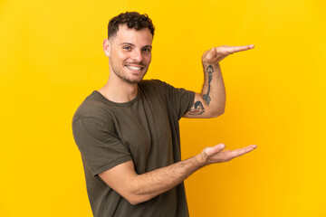 Young caucasian handsome man isolated on yellow background holding copyspace to insert an ad