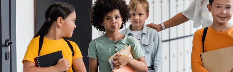 Obraz premium pensive african american schoolboy holding notebooks near multiethnic pupils and teacher, banner