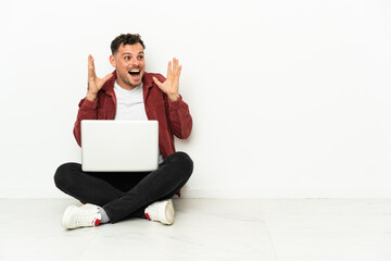 Young handsome caucasian man sit-in on the floor with laptop with surprise facial expression
