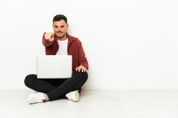 Young handsome caucasian man sit-in on the floor with laptop points finger at you with a confident expression