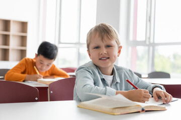 Fototapeta na wymiar happy schoolkid smiling at camera near asian boy writing on blurred background
