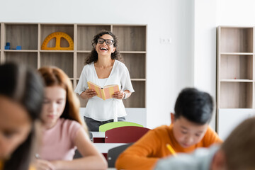 excited african american teacher holding book and laughing during lesson