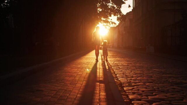 Silhouette of unknown mom and little daughter walking along the street paved with cobblestone in the evening
