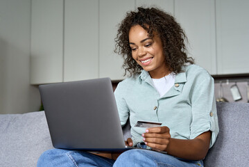 Young happy African American female consumer holding credit card and pc laptop sitting on sofa at home doing online banking transaction. Ecommerce virtual shopping, secure internet banking concept.