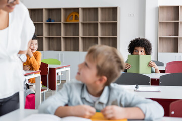 african american schoolboy holding book near blurred classmates and teacher