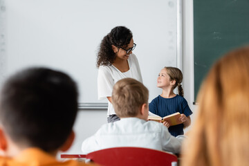 girl with book and positive african american teacher looking at each other while standing in classroom