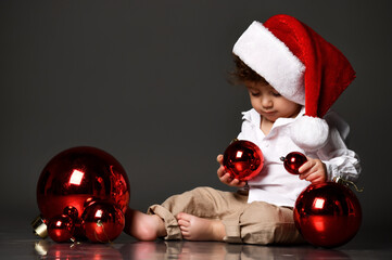 little boy in a santa hat plays on the floor with Christmas decorations balls. baby's first christmas, christmas eve