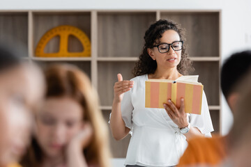 african american teacher reading book near blurred pupils in classroom