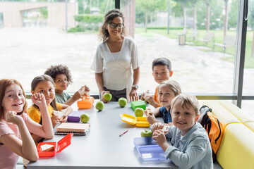 happy multiethnic schoolkids looking at camera near african american teacher in school eatery