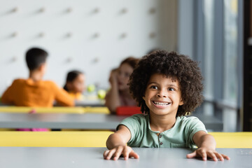 cheerful african american kid sitting in school eatery near pupils on blurred background