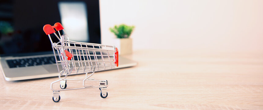 Close up the mini shopping cart and laptop at desk concept using the internet application for online shopping buying and spending on computer technology.