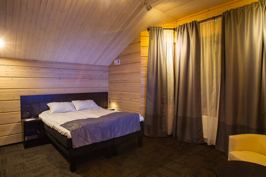 Attic Bedroom Interior. Concept - A Hotel Room. Hotel Room Is Located In Attic. A Double Bed Stands At Back. Double Bed With White Pastel Linen. Walls Of Bedroom Are Finished With Wood.