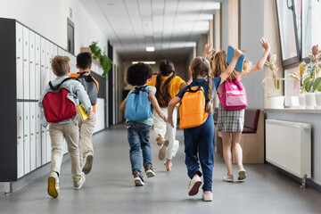 back view of interracial kids with backpacks waving hands while running in school hall