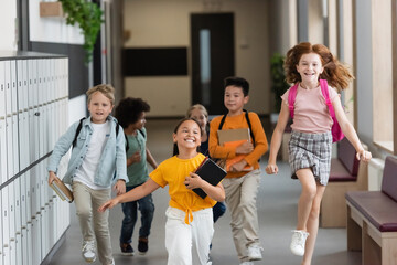 excited redhead girl jumping near multiethnic classmates running in school corridor