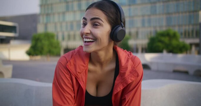 Positive young woman catches rhythm of music in headphones leads active lifestyle enjoys great morning has good mood talks with trainer sits on city background. Daily exercising to optimize health