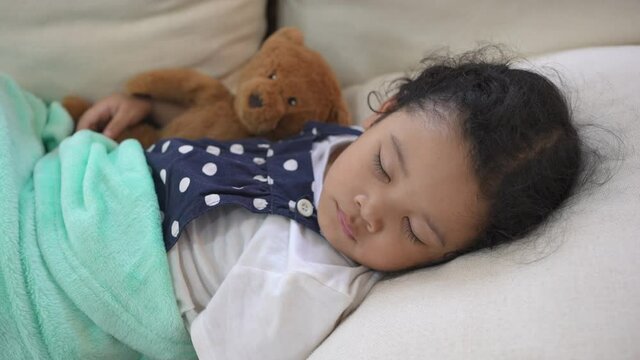 Adorable child girl kid sleeping on sofa in living room and hugging bear doll. Little cute daughter have day napping while mother do housework. Family bonding relationship and baby health care concept
