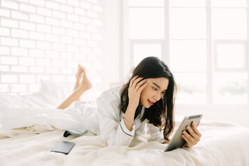 Obraz na płótnie Canvas A cute Asian woman with long black hair wearing white pajamas lying in a bed at home. A beautiful young woman waking up in the morning sunlight holding and using a tablet in her hand.