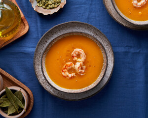 Autumn rustic homemade pumpkin and carrot cream puree soup with shrimps and condiment in bowl on blue linen tablecloth. Top view, flat lay, close up, horizontal. Concept of eat less meat food