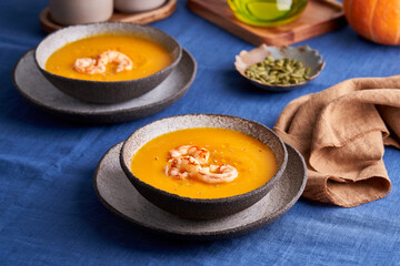 Healthy autumn pumpkin and carrot cream puree soup with prawn and seeds in bowl on blue linen tablecloth. Side view, close up, horizontal. Concept of eat less meat food