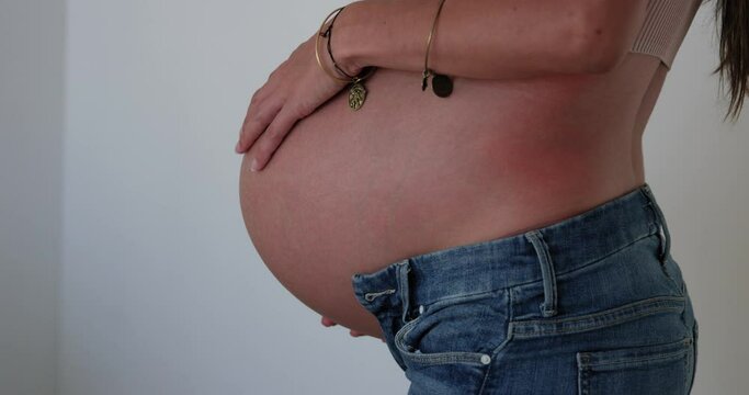 9 month pregnant woman rubs belly while wearing jeans against white wall - side profile