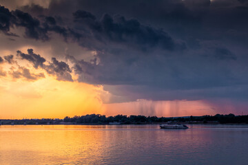 A small ship is sailing along the river against the backdrop of the sunset. Rain clouds in the sky. It's raining in the distance