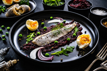 Marinated herring fillet with beetroots puree, hard boiled boiled egg, onion and green peas on wooden black table
