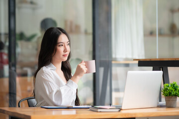 Photo of positive Asian woman holds a cup of coffee imagines plan holiday in office.