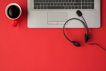 Top view photo of grey laptop red cup of coffee and black wired headphones with microphone on isolated red background with empty space