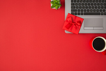 Top view photo of small vivid red giftbox with red ribbon bow on laptop plant and cup of drink on isolated red background with copyspace