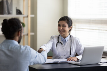 Happy young female indian ethnicity general practitioner physician therapist shaking hands with african american male patient, getting acquainted or making agreement at meeting in clinic office. - Powered by Adobe