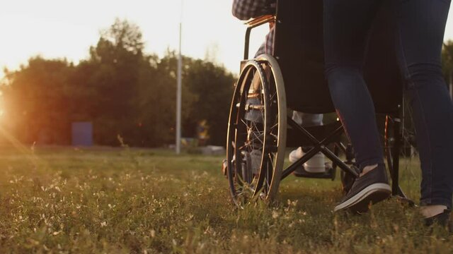 Woman rolls disabled person in wheelchair through field against background of sunset, back down view. Closeup walking legs. Concept supporting people with disabilities and overcoming any obstacles