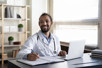 Distracted from computer and paperwork smiling pleasant african american doctor physician looking away, listening to colleague or thinking on pleasant news, sitting at table in clinic office.