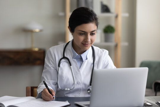 Concentrated millennial indian female gp doctor therapist working on computer, making notes in registration paper journal, prescribing illness treatment or consulting patient online in clinic office.