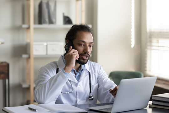 Concentrated young male african american general practitioner doctor holding distant phone call conversation working on computer in clinic office, giving distant professional healthcare consultation.