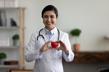Happy confident indian ethnicity female young doctor cardiologist holding heart figure in hands,...