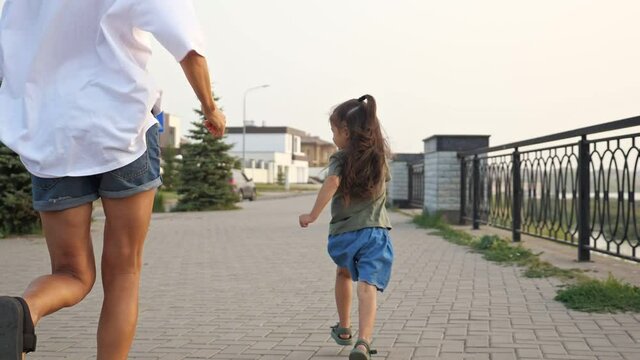Playful young woman in denim shorts and little daughter run along modern city embankment with railing at sunset time slow motion backside view