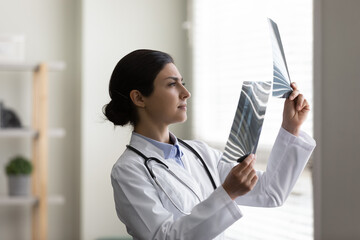 Concentrated young indian ethnic female surgeon traumatologist in white uniform looking at x-ray...