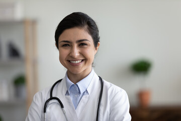 Head shot portrait of smiling confident young indian ethnicity female general practitioner therapist doctor in white coat posing in clinic room, professional healthcare medical services concept.
