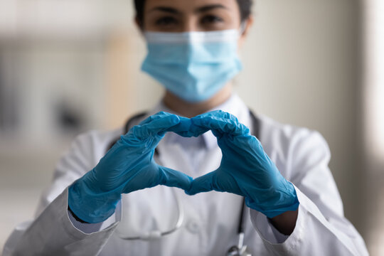 Close up millennial indian ethic medical worker general practitioner doctor in facemask and protective gloves showing love heart sign, expressing support to patient, charity donation concept.