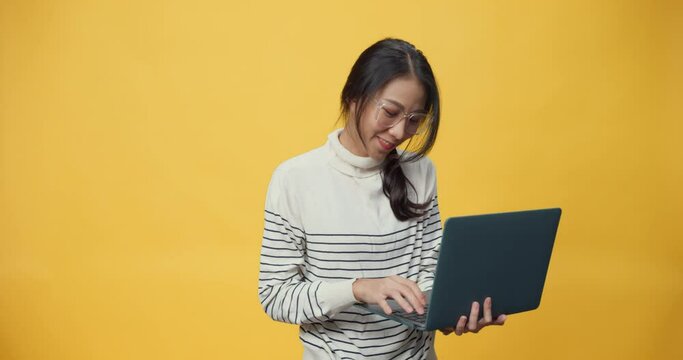Happy attractive Asia freelancer lady with casual cloth holding laptop computer and enjoy working dance singing and typing work at copy space while standing isolated over yellow background.