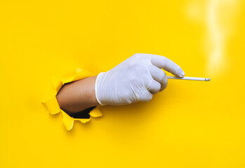 The hand of a doctor (nurse) in a white protective medical glove holds a slim cigarette.Torn hole in yellow paper. The concept of smoking, habit control among health workers during the covid-19 period