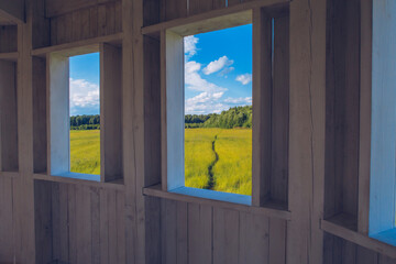 Path in the grass field through the wooden window view. Meadow picturesque summer landscape with clouds on blue marvelous sky background. Window portal to the nature landscape.