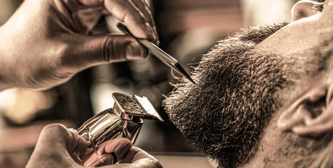 Barber works with a beard clipper. Hipster client getting haircut. Hands of a hairdresser with a beard clipper, closeup. Bearded man in barbershop. Man visiting hairstylist in barbershop - obrazy, fototapety, plakaty