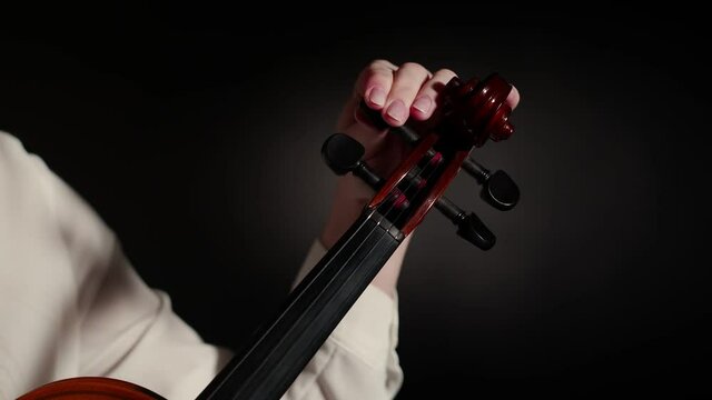 Violinist woman adjusts the violin, twisting pegs with hand on a dark studio background, close-up. Musician adjusts strings of violin, rotating the pegs. Concept of a string musical instrument.