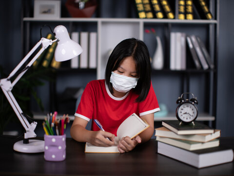 school girl pupil wearing the mask studying at home sitting at desk. Smart cute kid primary school student writing in exercise book doing homework, learning at table.