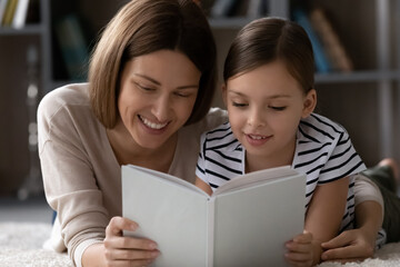 Happy bonding young mother and little kid daughter reading interesting fairy tale story in paper book, enjoying domestic family hobby activity pastime together lying on floor carpet in living room.