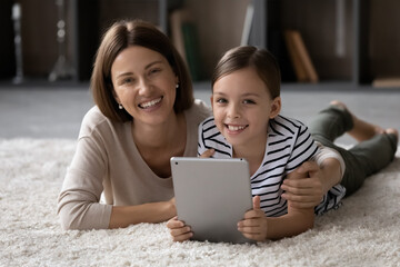 Portrait of loving affectionate young mother cuddling little cute kid daughter, using digital touchpad together, lying on cozy fluffy carpet in modern living room, happy female family weekend pastime.