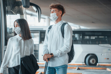 Jóvenes adolescentes con mascara en la estación de autobuses esperando su siguiente parada con equipaje y mochila - obrazy, fototapety, plakaty